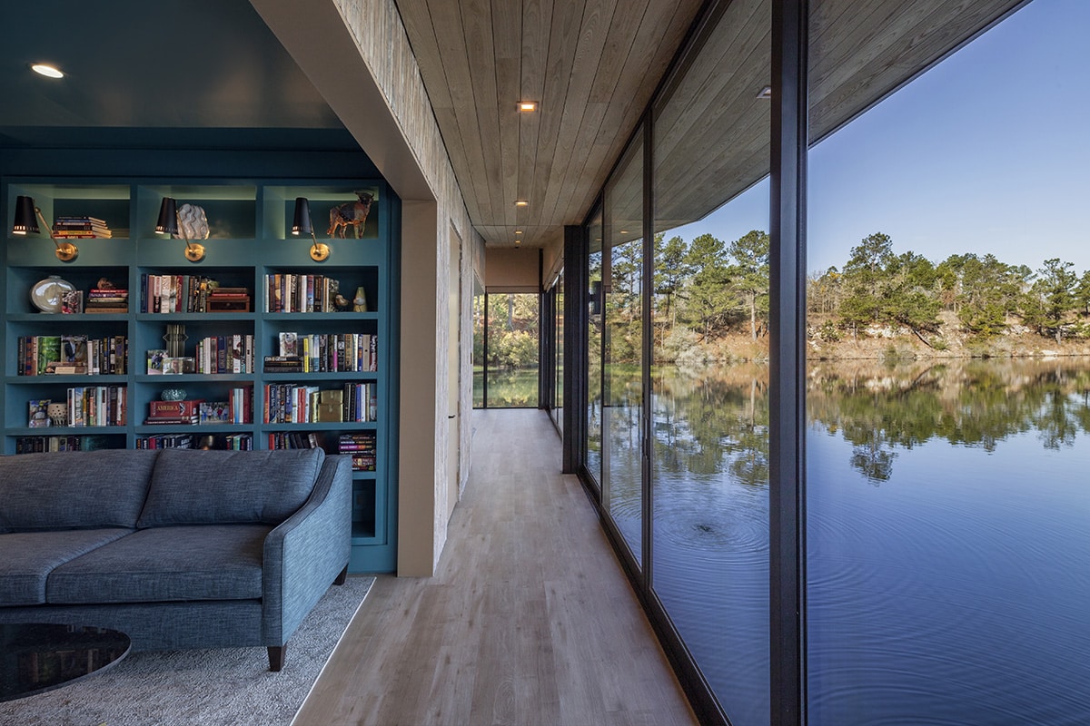 Photo of the living room of a house floating on a lake. The right side is a floor to ceiling wall of glass panels with black aluminum frames, with a perfect view of the lake and distant shore. 