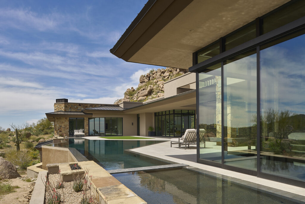 Outdoor shot of a modern desert house from the polygonal pool. The house is lined with large windows and multi sliding doors with black aluminum frames that reach from floor to ceiling. 